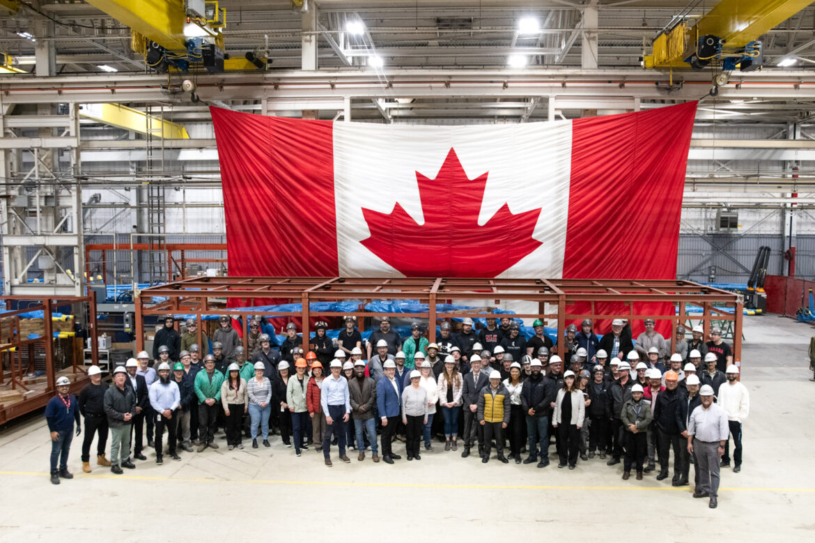 People stand in front of feeders and large Canadian Flag at BWXT in Cambridge.