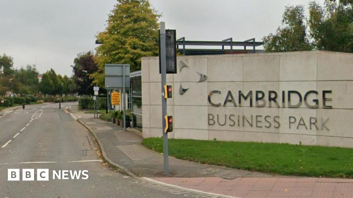 The entrance to the Cambridge Business Park. On the right is a stone wall with Cambridge Business Park written on it in silver letters. There is a traffic light in front of it. The road stretches down and has small trees. 