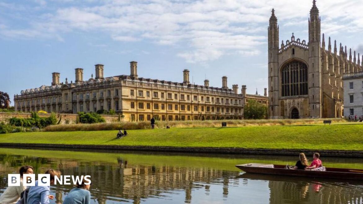 Three students sit in the foreground by the river Cam, opposite Kings College Cambridge on a sunny day. Two more students are in a punt on the river passing by in front of them. 