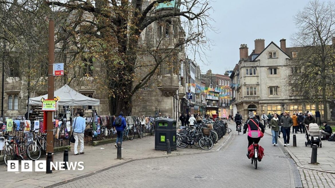 Looking down a street in Cambridge city centre. Two cyclists are riding away from the camera. Black iron railings and a row of parked bikes are to the left, and a terrace of old buildings curves to the right in the distance. Groups of pedestrians walk on the paths either side.  
