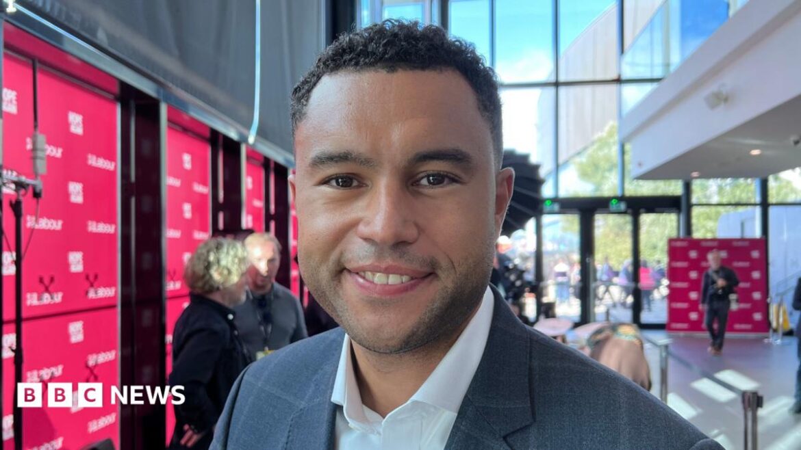 Man with curly black hair wearing a suit stands in front of Labour branding.